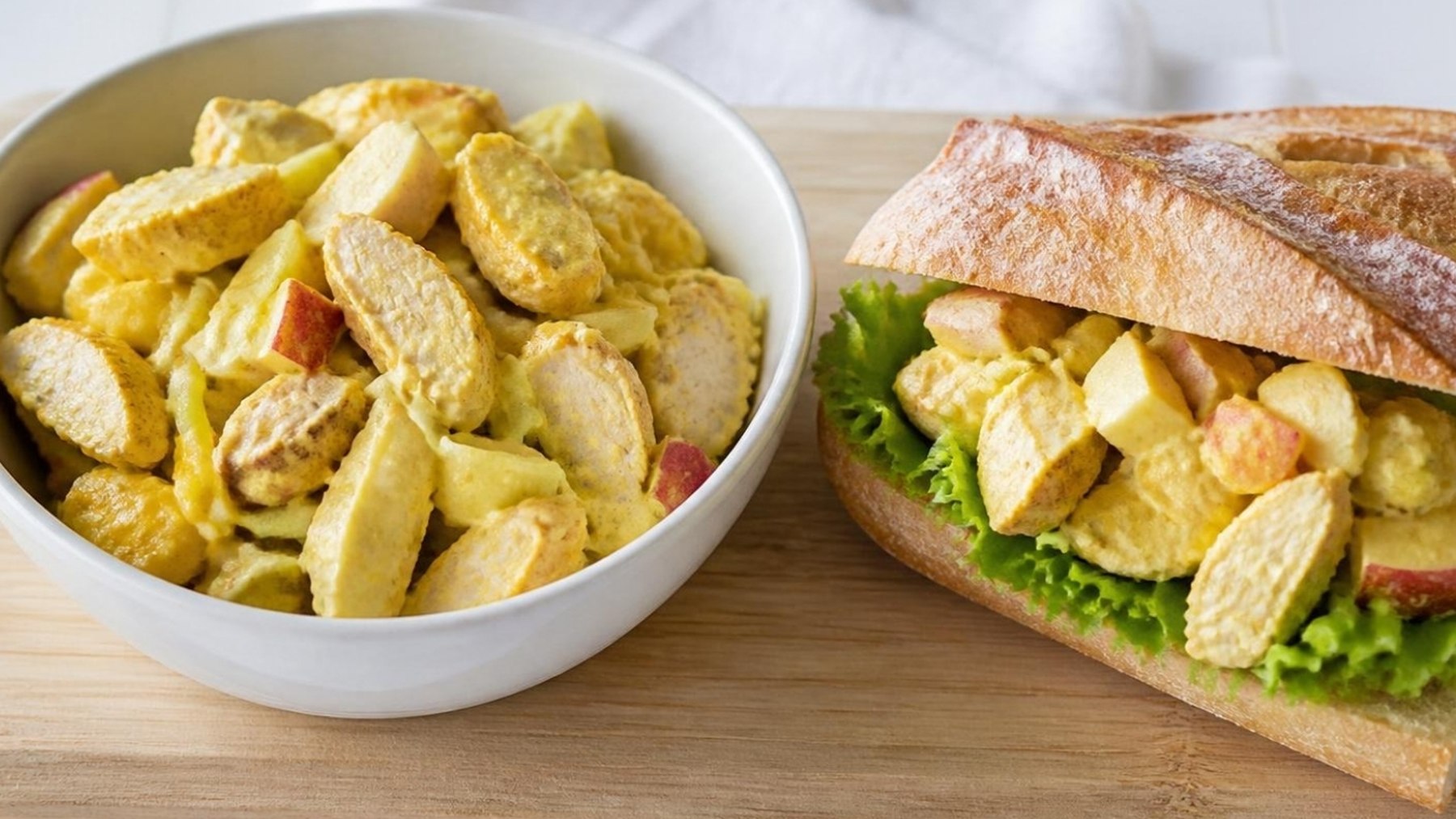 Kall kycklingcurryröra med äpple i en skål, bredvid en baguette fylld med röran och grönsallad