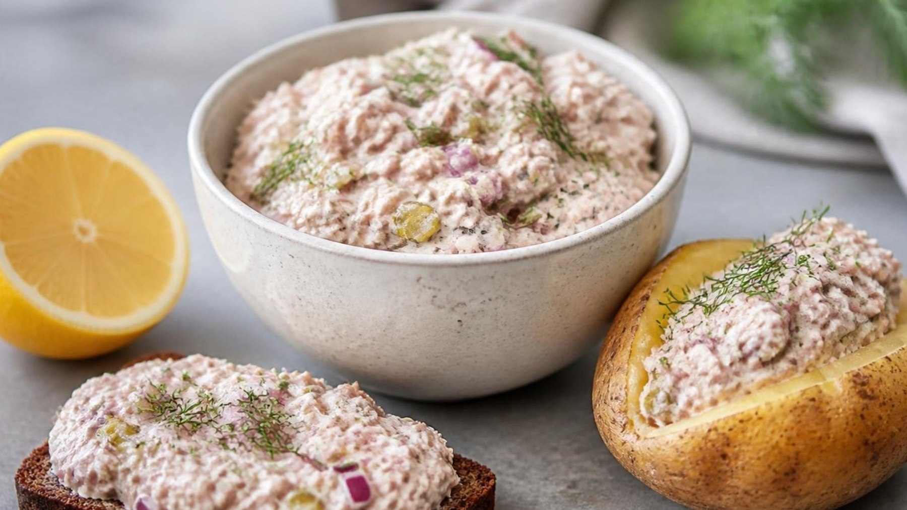 Tonfiskröra med rödlök och dill i en skål, bredvid en kluven bakad potatis, en smörgås med röran och en halv citron
